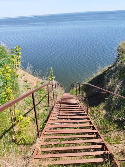 The image shows a rusty metal staircase descending towards a body of water. The location is likely in Икское Устье, Russia, as provided. The staircase has simple railings on either side, and the surrounding terrain is grassy. The water is calm with small ripples. The sky is clear and blue. There are no people visible in the frame.