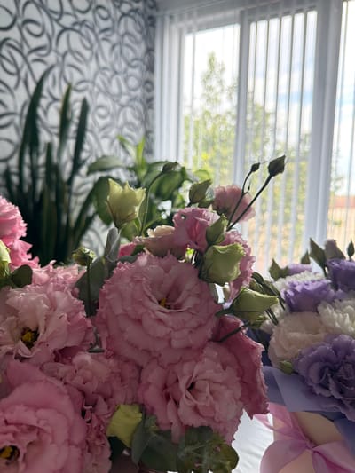 A bouquet of pink, white, and purple lisianthus flowers is arranged on a table in front of a window with vertical blinds. The flowers are predominantly pink, with some buds showing green. Behind the flowers, a tall green plant with long, sword-like leaves is visible against a wall with a swirling grey and white pattern. The background outside the window is blurred, showing trees and a building. The scene suggests an indoor setting, possibly a home or a florist's shop. The location is identified as Ust'-Dzhigutinka, Russia.