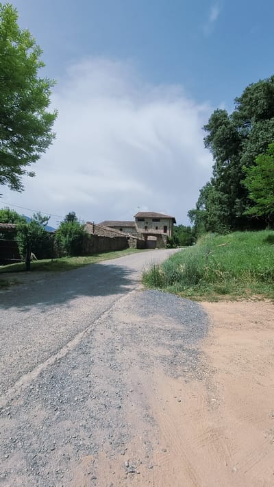 The image depicts a rural scene in la Vall d'en Bas, Spain, under a partly cloudy sky. A dirt road, with tire tracks visible, occupies the right side of the frame, curving towards the background. The road is bordered by a grassy verge.
To the left of the road, a rustic stone building with a tiled roof is visible. It appears to be an old farmhouse or a rural dwelling, with multiple windows and an arched entryway. Green trees and foliage surround the building and extend into the distance, suggesting a natural, possibly wooded, environment.
The time of day appears to be daytime, with soft shadows cast on the road, indicating the sun is not directly overhead. The weather seems pleasant, with a mix of blue sky and white clouds. The overall mood of the image is peaceful and serene, evoking a sense of rural tranquility. There is no visible text in the image.