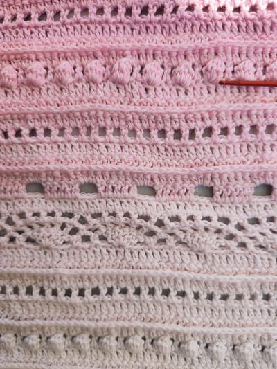The image shows a close-up, overhead view of a crocheted fabric with a gradient of pink and white colors. The fabric features several distinct rows of stitches, including rows of bobble stitches, solid crocheted rows, and openwork or lacy rows. A red crochet hook is visible on the right side of the image, resting on the crocheted fabric. The lighting suggests an indoor setting. The image does not contain any information that would allow for identification of its location in Durlești, Moldova, or any details about people, actions, or interactions.