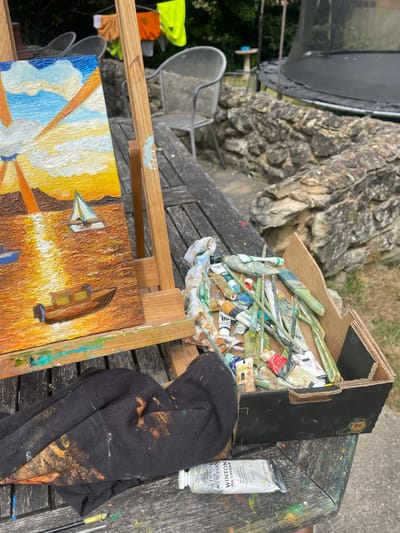The image shows an artist's outdoor painting setup, likely in a garden. A wooden easel holds a painting depicting boats with a warm-toned, impressionistic style. A cardboard box filled with paint tubes and brushes sits on a weathered wooden table. A black cloth splattered with paint rests near a tube of Winsor & Newton Winton oil paint. In the background are stone features and a trampoline, suggesting a residential area. The label on the paint tube indicates "Winton Oil Colour". There's no visible indication of the location being in Maidstone, United Kingdom.