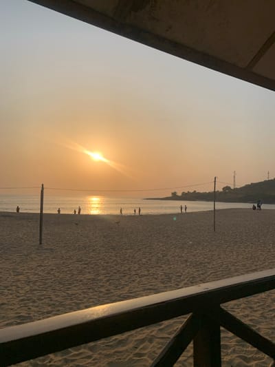 The image captures a serene sunset or sunrise scene on a beach, likely in Goderich, Sierra Leone, given the context. The photograph is oriented in landscape format, with a warm, golden-orange hue dominating the sky and water.
The sun, a bright glowing orb with prominent lens flare, is positioned towards the lower left, casting a shimmering path of light across the calm water. The sky, especially above the horizon, displays a soft gradient of light orange and yellow, transitioning to a paler hue higher up, suggesting a hazy atmosphere.
Along the waterline, several small, silhouetted human figures are visible, some appearing to be walking and others standing. Their presence adds a sense of scale and quiet activity to the vast expanse of sand and sea. The sandy beach occupies the mid-ground and right side of the frame, showing faint textures and possible footprints.
On the left and right sides, dark, angular structures frame the scene. On the far left, a solid, diagonal barrier or wall is visible, while on the far right, a dark, angular railing or framework extends into the sand. Two dark, slender poles stand vertically in the shallow water or sand, adding depth to the composition. In the distant background across the water, a faint outline of land or vegetation can be discerned. There is no visible text in the image. The overall impression is one of peaceful twilight on a coastal landscape.