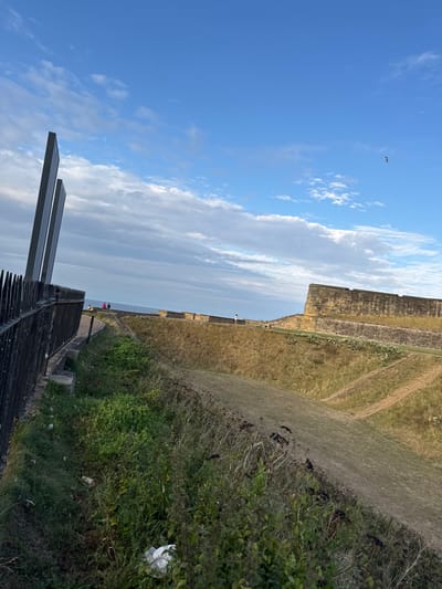 The image depicts an outdoor coastal scene in North Tyneside, United Kingdom, specifically near historical fortifications consistent with Tynemouth Priory and Castle.
In the foreground, a black metal fence with pointed vertical finials extends diagonally upwards from the bottom-left corner. Immediately behind the fence, a strip of unkempt vegetation, primarily green and brown weeds, contains a visible piece of white plastic litter.
The midground features a wide, dry, grassy ditch or moat-like depression with sloped banks. The grass on these banks is predominantly light brown and dried, with some greener patches. On the elevated far side of this depression, a fortified stone wall, constructed from light brown/sand-colored blocks, runs horizontally. This wall has a stepped base and a crenellated upper section. A winding path is visible on the upper bank, adjacent to the wall and overlooking the sea.
Four individuals are present on this elevated path. Two people are positioned together on the left, near the edge overlooking the sea, possibly seated or standing. A third person stands further right along the path, facing away from the viewer. A fourth individual is partially visible near the wall further right.
To the left of the image, rising behind the foreground fence, are two tall, slender, dark grey, rectangular monolithic structures. These appear to be part of a modern memorial or artistic installation.
The background consists of the North Sea horizon, appearing calm with a grey-blue surface. Above the sea, the sky displays patches of clear blue interspersed with scattered white and grey stratocumulus clouds. A single dark bird is in flight in the upper right quadrant of the sky. The scene is illuminated by natural daylight, indicating clear visibility.
