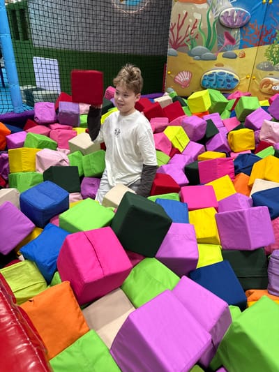 A boy is surrounded by colorful foam blocks in an indoor playground, with a wall mural depicting underwater sea life visible in the background. The playground is likely located in the Перовское сельское поселение, Ukraine.