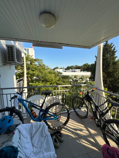 The image depicts a balcony in Obrochishte, Bulgaria, with two mountain bikes parked side-by-side. A blue Canyon mountain bike is on the left, and a dark-colored mountain bike is on the right. Several items are scattered on the balcony floor, including a white laundry basket filled with towels, a blue bicycle helmet, a pair of athletic shoes, and a pink bag. An air conditioning unit is mounted on the wall to the left, and a white plastic chair is positioned behind the blue bicycle. The balcony overlooks a lush green area with trees and buildings visible in the background. The sky is clear and blue, indicating a sunny day. The lighting suggests it is late afternoon due to the long shadows cast on the tiled balcony floor.