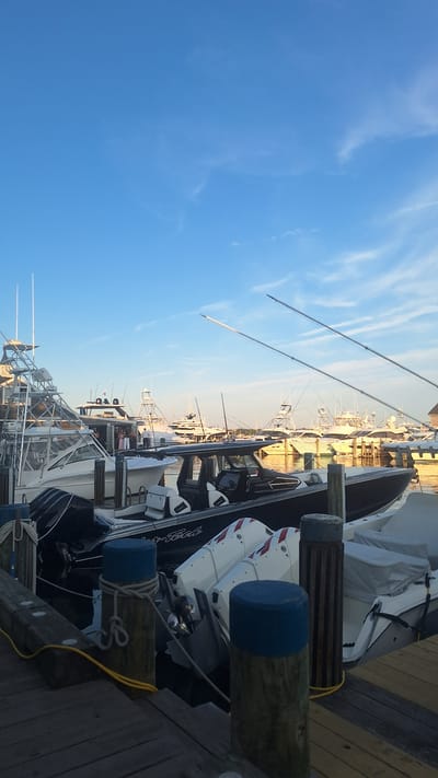 A view of a harbor in Nantucket, Massachusetts, United States of America, shows several boats docked at a pier. The sky is a clear blue with sparse white clouds. In the foreground, a wooden pier is visible, with three blue-capped bollards. A black boat with white seats and twin outboard motors is docked to the right. To the left of this boat, a white cabin cruiser is also docked, with people visible on deck. In the background, a line of larger yachts and fishing boats are moored. The sun is low in the sky, casting a warm light on the water and boats. Fishing rods extend from some of the boats.