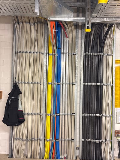The image shows a section of a utility room with a large number of organized electrical cables. The cables are bundled and secured with metal clamps to a metal frame attached to a brick wall. There are multiple rows of cables, predominantly beige and black, with some colorful cables (yellow, orange, blue, red) visible in the center. A black jacket is hanging on the left side of the cable bundles. Above the cables, metal conduit and cable trays are visible. On the right side of the image, a yellow sign with German text is mounted on the wall, and further down, electrical junction boxes and connectors are present. The overall environment suggests a technical or industrial setting. The location of Mannheim, Germany is not discernible from the image itself.