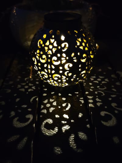 The image shows a close-up, low-angle, dimly lit shot of a decorative, spherical lantern with intricate cutouts. The lantern is dark in color and appears to be made of metal. Warm yellow light emanates from within the lantern, casting decorative patterns of light and shadow onto the dark, textured surface it rests upon. The surface appears to be a wooden table or deck. The background is dark and out of focus, with some faint reflections visible in the upper left portion of the image. There are no people visible in the image, nor any signs of the city of Pyatigorsk, Russia.