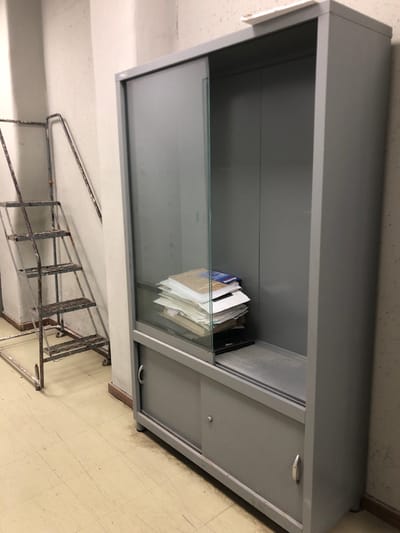 The image shows a gray metal cabinet with a glass sliding door on the top half and two solid sliding doors on the bottom half. The cabinet is located in a room with a beige floor, an off-white wall, and a metal ladder against the wall to the left of the cabinet. There are a stack of papers on the top shelf of the cabinet, behind the glass sliding door. The cabinet appears to be empty otherwise. The coordinates provided place the image in Agia Paraskevi, Greece. The cabinet is located in a room with a beige floor.