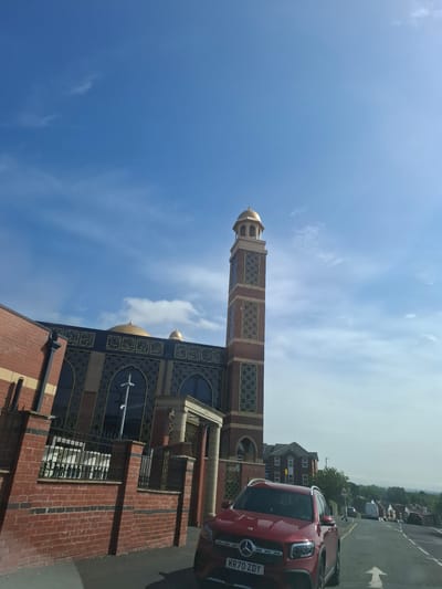 A wide shot captures the exterior of a mosque in Manchester, United Kingdom, on a bright, sunny day with a blue sky dotted with sparse white clouds. The mosque features a tall, brick minaret topped with a golden dome, and a main building with a large golden dome. The architecture incorporates intricate golden Islamic calligraphy and geometric patterns against a dark facade, with large arched windows. In the foreground, a red Mercedes-Benz SUV is parked on the right side of the road. The vehicle has the license plate KR70 ZDY. A red brick wall with a black metal fence lines the front of the mosque. To the right of the mosque, a residential street with houses and trees extends into the distance. The road has white lane markings and a pedestrian crossing.