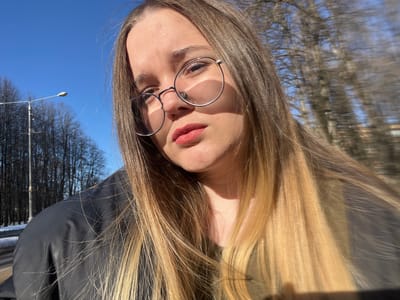 A young adult female is depicted in a close-up, selfie-style portrait taken outdoors in Pushchino, Russia. She wears round, metallic-framed glasses and reddish-pink lipstick, gazing directly into the camera. Her long, straight, light-colored hair, transitioning from brown at the roots to blonde ends, frames her face and extends over her dark-colored jacket and visible olive-green undershirt. Bright sunlight illuminates her from the left, creating strong shadows from her glasses on her face. The background, largely blurred, shows a clear blue sky above a line of leafless trees, consistent with late autumn or winter conditions. A street lamp on a tall pole is visible to the left, adjacent to a paved area with remnants of snow on the ground.