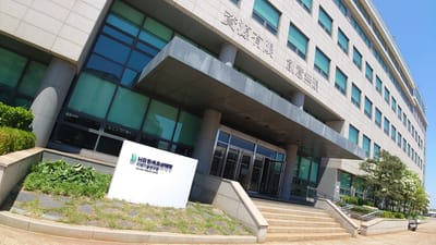 The image shows the exterior of a multi-story building, likely an office or research facility, located in Ulsan, South Korea. The building is constructed with light-colored stone or concrete panels and features rows of windows with teal-colored blinds. Above the entrance, there is a protruding, gray overhang supported by cylindrical columns.
In front of the building, a white sign is visible with Korean and English text, identifying the location as the "HD 한국조선해양 미래기술연구원 / HD Korea Shipbuilding & Offshore Engineering Advanced Research Center."
The entrance is accessible via a set of stairs. Greenery, including bushes, grass, and trees, surrounds the building. The sky is blue. A few vehicles are visible in the far right background. No people are visible in the image.