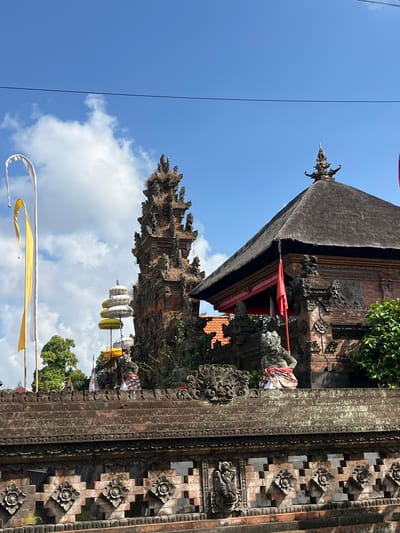 A temple complex is captured from a low-angle exterior view on a sunny day in Abiansemal, Indonesia. The foreground features a detailed stone wall with intricate carvings of floral patterns and mythological figures. Behind the wall, a multi-tiered pagoda-like structure is visible, adorned with yellow and white layered umbrellas. To the right, a larger temple building with a dark, thatched roof and brick walls stands prominently. Red and white flags are displayed on poles, and a single red flag is also attached to the main temple building. Two stone statues, one appearing to be a guardian figure with a patterned cloth around its waist, are positioned on the upper level of the temple wall. The sky above is bright blue with scattered white clouds. Tall trees and other temple structures are visible in the background.