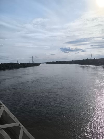 An overcast, daytime view of a wide, dark river, likely the Volga River near Kuz'minka, Russia. The water is choppy, reflecting the dull sky and distant sunlight. The riverbanks are lined with dense, dark green trees. Two tall, lattice-work power line towers are visible on the left bank. On the right bank, the tree line continues, with a hint of a distant structure. The foreground shows a portion of a grey metal bridge structure, with visible rivets and crossbeams. The overall scene is one of natural landscape and industrial infrastructure under a muted sky.