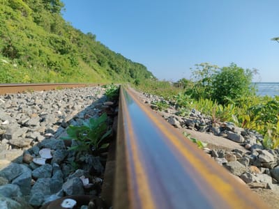 The image shows a close-up view of a rusty railroad track running along a steep, tree-covered embankment. The tracks are surrounded by grey gravel and some green plants are growing between the stones. The scene is bathed in sunlight under a clear blue sky. The location is Tret'ya Pad', Russia. In the background, the sea can be seen.