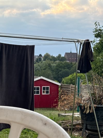 The image depicts a cloudy sky over a suburban landscape. In the foreground, a white plastic chair is partially visible, with a clothesline stretching horizontally across the frame, holding a dark fabric and a dark jacket. To the right of the clothesline, a green leafy tree branches into the frame. In the middle ground, a small, red shed with white-framed windows is situated, with a glimpse of something within one of the windows. Behind the shed, a wooden fence and dry, brown vegetation are visible. In the background, a dense forest covers a hillside, and further in the distance, a large, brick building with a pointed spire, likely a church, is nestled amongst the trees. The overall impression is of a residential area with a church steeple visible in the distance, possibly in or near the city of Stolberg, Germany, given the architectural style of the church.