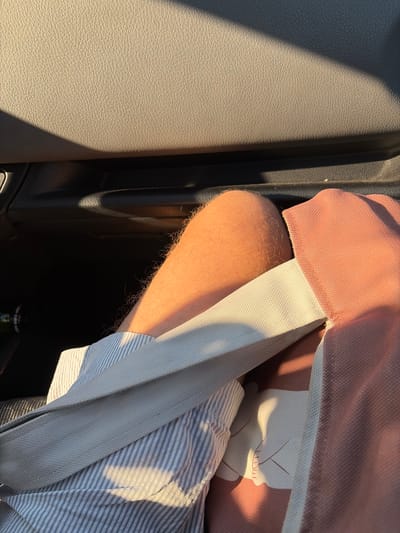 The image shows a close-up of a person's arm and shoulder in a car. The person is wearing a pink shirt and a striped blue and white shirt underneath. A light-colored seatbelt crosses over the person's shoulder and chest. The arm is hairy and illuminated by sunlight, suggesting it is daytime. Shadows indicate the light source is coming from the right, casting long shadows across the scene.
The background of the image shows the interior of a car, with dark gray textured plastic on the left and a black void that appears to be a storage compartment or the space between seats on the right. A hint of a green bottle is visible at the bottom right corner, possibly a beverage. The overall impression is of a person sitting in a vehicle, perhaps on a journey. The warm sunlight suggests it could be late afternoon or early morning. No text is visible in the image. The scene is set in Škaljari, Montenegro.