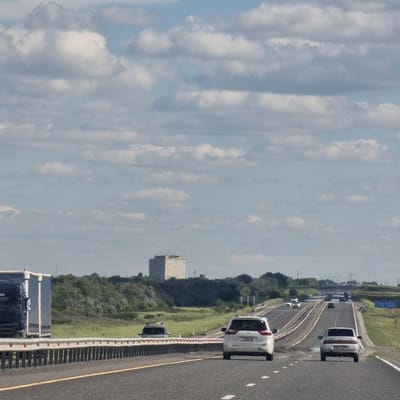 The image depicts a highway scene, likely taken from a moving vehicle. Several cars, including a white SUV and a dark sedan, are visible on the multi-lane asphalt road. The road has yellow and white lane markings. On the left side of the road, there is a grassy embankment with trees and a large, light-colored rectangular building in the distance. The sky is partly cloudy with streaks of white clouds against a pale blue background. The overall impression is of a daytime scene, possibly mid-morning or afternoon, with fair weather. A blue sign with white text is visible on the side of the road in the upper left portion of the image, though the text is not legible. The image is oriented vertically, suggesting it may have been rotated.