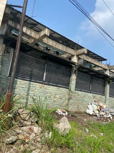 The image shows a concrete wall with a black metal fence and a stone facade. The wall is located on a sloping lot, and the ground is covered in grass and rubble. There are several white bags and other debris lying on the ground. The scene is taken from a low angle, looking up at the wall. The coordinates provided indicate a location in the city of Margaasih, Indonesia, which is about 10 kilometers south of Bandung, the capital of West Java.