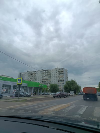 The image depicts a street scene in Orekhovo-Zuyevo, Russia, under a cloudy, overcast sky, suggesting it is daytime. The foreground is dominated by the interior of a vehicle, likely the viewpoint of someone driving or a passenger. Beyond the vehicle, a wide asphalt road is visible, with a pedestrian crossing marked in yellow and white stripes.
On the left side of the road, a modern green and white supermarket building is present, with large glass windows and signage. The text "Пятёрочка" (Pyaterochka) is visible on the building, indicating it is a well-known Russian retail chain. There are also price signs visible through the windows, with numbers like "79" and "30". A pedestrian crossing sign, a yellow triangle with a blue pedestrian symbol, is prominently displayed near the supermarket entrance. A few people are seen on the sidewalk in front of the store, some carrying bags. Several cars are parked along the sidewalk and on the street.
Across the street from the supermarket, a multi-story residential building stands, typical of Soviet-era architecture. It is a large, grey concrete structure with numerous windows. Further down the street, trees and more buildings are visible. In the right lane of the road, a large orange tanker truck is driving away from the camera. Another pedestrian crossing is visible in the distance, and more vehicles are present on the road. The overall atmosphere is that of a typical urban environment on a slightly gloomy day.