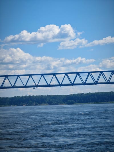 The image captures a wide-angle view of a blue metal truss bridge spanning a broad, dark blue river under a partly cloudy sky. The bridge's geometric structure is prominent, with triangular supports forming a repeating pattern across its length. In the distance, a dense, dark green treeline borders the far bank of the river, suggesting a natural landscape. Faint white structures, possibly buildings or remnants of a dock, are visible along the shoreline. The water's surface is rippled, indicating a gentle current or breeze, and a few small white buoys or markers are scattered in the river. The sky is a vibrant blue, dotted with fluffy white clouds. The overall scene depicts a tranquil day, likely during daylight hours, with the focus on the impressive man-made structure against a natural backdrop. The image does not contain any people or discernable activity.