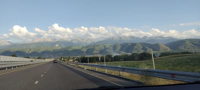 The image captures a forward-facing perspective from a vehicle driving on a multi-lane highway that cuts through a landscape with distant mountains. The highway, paved with asphalt, has clear lane markings and is bordered by a metal guardrail. To the left, a sound barrier wall runs along the highway. On the right side of the road, a grassy field slopes upwards towards a range of green, undulating mountains. Some scattered houses and trees are visible in the foothills and along the base of the mountains. The sky is partly cloudy with large, white cumulus clouds scattered against a light blue backdrop. Two vehicles are visible on the highway; a dark-colored SUV in the distance on the right lane, and another dark vehicle further ahead on the left lane. The overall scene suggests a drive through a semi-rural or suburban area leading towards a mountainous region. The location is identified as Қырғауылды, Kazakhstan.