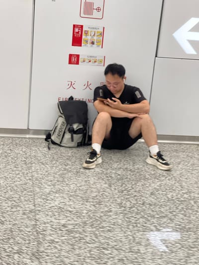 A young Asian man wearing glasses, a black t-shirt, and black shorts is sitting on the floor in what appears to be an indoor public space, possibly an airport terminal or train station, in Panyu District, China. He is looking down at his phone, which he holds in his hands. The man has short black hair and is wearing white earbuds. Beside him is a gray and black backpack with the word "THERONTUR" printed on it. Behind the man is a white wall with several signs, including a red sign with Chinese characters and pictograms related to fire safety. One sign has the words "FIRE EXTINGUISHER" printed in English. To the right of the man, on the wall, is a large white arrow pointing to the left, indicating direction. The floor is tiled with a speckled gray pattern.
