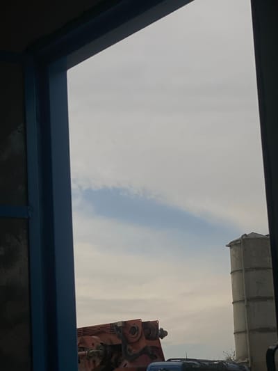 A blue-framed rectangular opening reveals a sky filled with light gray clouds, with a vertical strip of light blue sky cutting through them. Partial views of industrial structures, possibly construction-related, appear at the frame's right edge, including what looks like the top of a silo and mechanical equipment. The bottom portion of the frame shows reflective surfaces.
