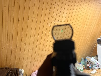 The image depicts a first-person perspective through a rifle scope, targeting a wooden wall. A green crosshair reticle is visible within the scope's view. In the background, to the right, are various objects including a white refrigerator, a microwave, and a collection of plastic bottles and other miscellaneous items. The wall is constructed of vertical wooden planks. The scene appears to be an indoor setting, possibly a room. There is no discernible information in the image to indicate the location is Vasilyevo, Russia, nor is there any evidence of people or specific actions taking place.