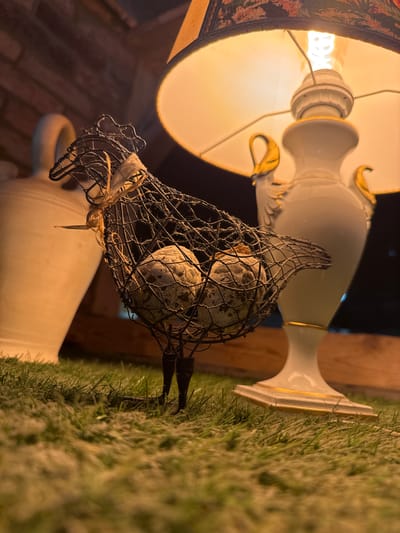 The image depicts a dimly lit still life, possibly an indoor scene given the presence of a lamp and a ceiling structure. The primary focus is a decorative wire birdcage filled with speckled decorative eggs, resting on a white ceramic lamp base adorned with gold accents. The lamp is turned on, casting a warm, orange glow through its patterned lampshade. To the left, a patch of artificial grass is visible, its texture slightly blurred. In the background, hints of wooden beams and a large white vase can be seen. The overall mood is cozy and perhaps a bit whimsical, suggested by the decorative elements. The lighting indicates it could be nighttime or a dimly lit indoor space. No people, text, or weather conditions are apparent. The context provided suggests a location in Sankt Wendel, Germany.