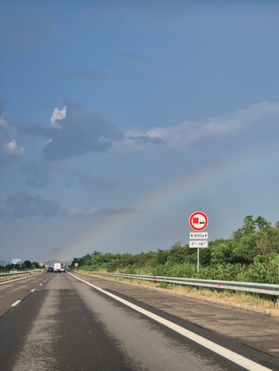 This media file captures a view of a highway in Krustina, Bulgaria, under a sky with scattered clouds and a faint rainbow. The road appears wet, suggesting recent rain or high humidity, and the scene is set during the daytime.

The primary focus is the highway itself, stretching into the distance with a white line marking the lanes. Guardrails line the left side of the road, separating it from a dense line of green trees and vegetation. A traffic sign is visible on a pole, indicating "↑ 4000M" and a time frame of "7⁰⁰-19⁰⁰," suggesting a restricted zone or a specific route for certain vehicles. Another speed limit sign, marked with "60," is visible further down the road.

Several vehicles, including cars and what appear to be vans or trucks, are traveling on the highway, contributing to the sense of ongoing activity. The sky transitions from a bright blue in the upper left to a more overcast, grayish hue towards the right, where the rainbow is faintly visible. The overall mood is calm and serene, with a touch of natural beauty provided by the rainbow and lush greenery. The location context of Krustina, Bulgaria, places the scene within a specific geographical setting.