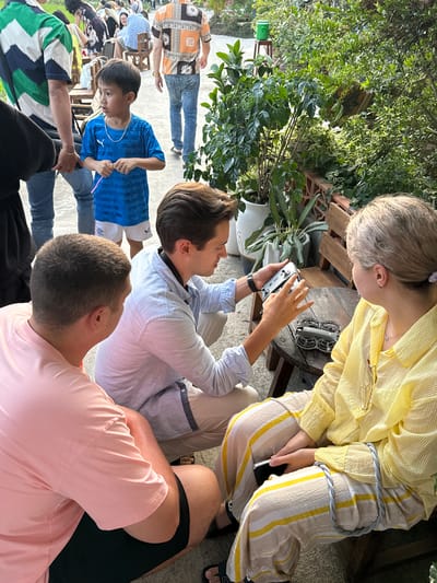 This media file captures an outdoor gathering in Diên Khánh, Vietnam, bathed in bright daylight. The scene features multiple individuals interacting in a lush, green environment.
In the central foreground, a person with dark hair, wearing a light blue striped long-sleeve shirt and light trousers, is intently focused on a white drone controller they are holding. Beside them, a person with short, light-colored hair, dressed in a yellow striped long-sleeve shirt and matching striped trousers, leans in, observing the controller with interest. A small, gray drone with four propellers is placed on a rustic wooden table next to them. The person in yellow also holds a smartphone in their lap, on the upper thigh area of the right leg, a small "FILA" logo is visible on the striped pants.
To the right, the back of a head and shoulder of a person wearing a pink t-shirt are visible. Further back, a young boy in a blue t-shirt and dark pants stands, looking towards the drone and its handlers. Several other people are partially visible in the background, including one person in an orange and black patterned shirt, suggesting a larger social event.
The setting is an outdoor space, likely a garden or patio, characterized by an abundance of green foliage, potted plants, and wooden furniture. The ground appears to be paved. The overall mood is one of engaged curiosity and social interaction, centered around the drone.