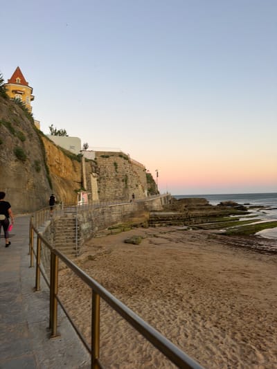 The image captures a view along a coastal path in Cascais, Portugal, during what appears to be late afternoon or early evening. A paved walkway with a metal railing runs along the left side of the frame, bordered by a steep, rocky embankment covered in sparse vegetation. Atop the embankment, a distinctive yellow building with a red, pointed roof is visible. Further along the path, stone steps lead down towards a sandy beach.
The beach itself is composed of light brown sand, with some scattered rocks and areas of seaweed. The sea is to the right, with a rocky coastline jutting out into the water. The water appears calm, and the sky above is a clear gradient of light blue to a soft pinkish hue near the horizon, indicating sunset.
There are a few individuals on the path. In the foreground, a woman with dark hair wearing dark athletic wear and carrying a pink bag walks away from the camera. Further up the path, a person in dark clothing is running. Another person is visible walking further in the distance on the path. The scene suggests a leisurely or recreational activity taking place by the coast.