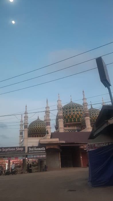 An evening street scene in RawangItiek, Indonesia, captured at eye-level, features a large mosque and adjacent commercial structures under a light blue sky. Two distinct bright light sources are visible in the upper left sky. Multiple horizontal utility lines traverse the mid-upper frame.

The central background is dominated by a mosque, characterized by multiple white minarets with pointed finials and at least three prominent domes adorned with intricate green and gold geometric patterns. White building facades with arched decorative elements are visible beneath the domes.

In the foreground, a rough ground surface suggests an unpaved road or dirt path. On the left, two adjacent commercial storefronts are visible. The first, labeled "Dea Septiana Shop," displays merchandise within. A person in a white shirt and dark pants stands beside a light-colored motorbike parked facing right near this shop. Another individual is partially visible inside. The second shop, "Desi Kosmetik," also displays products, with two individuals in dark clothing visible inside and another person seated outside to the right.

To the far right, a structure is partially obscured by a dark blue tarp. A sign above it reads "RUMAH BA...", with the full text obscured. A dark-roofed building, possibly connected to or adjacent to the mosque complex, stands between the commercial shops and the tarp-covered structure. A dark, rectangular street lamp fixture is partially visible on the far right.