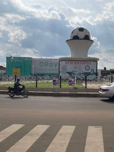 The image depicts a street scene in Sardauna, Nigeria. A large white structure topped with a black and white soccer ball dominates the center of the frame. A billboard is attached to the structure, displaying "NEW KASUPDA" and images of men in traditional attire. To the left, a green building with "OPay" signage is visible. In the foreground, a man is riding a motorcycle on the road. A white car is parked on the right side of the image. The sky is partly cloudy.