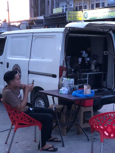This media file shows a street-side coffee stand operating out of the back of a white van. The van is parked on a paved surface, likely a street or sidewalk, with buildings and signage visible in the background. Two red plastic chairs and a few other chairs are set up for customers.
A young man is seated in one of the red chairs, looking at his phone. He is wearing a brown t-shirt and black pants. He appears relaxed. The coffee stand itself is equipped with various containers and supplies, including a blue plastic bag, suggesting it is open for business. There is a small table laden with items like cups and bottles.
Several signs are visible, some in Indonesian. One sign on the van reads "KOPI ZENA" and another says "IDRUZE OBRAL Paste Murak". Other signs in the background indicate commercial establishments, with text like "Rena Ulshop 2" and "DAFFA". The lighting suggests it is late afternoon or early evening. The overall scene depicts a casual outdoor setting for a food and beverage vendor.