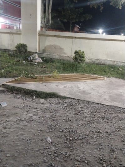 This image depicts an outdoor scene at night, likely in Keudeteupinpunti, Indonesia, given the location context. The setting appears to be an area with a concrete pathway and a grassy patch, bordered by a textured wall. In the foreground, a gritty, gravel-covered ground stretches out.
The central object of interest is a long, triangular structure composed of parallel wooden slats, possibly a decorative garden bed or a drainage grate, with some sparse vegetation growing within it. To the left of this structure, a small, bushy green plant is visible. Behind the grassy area, a light-colored wall with some discoloration and staining runs across the frame. A concrete pillar stands to the left of the wall. In the background, trees are silhouetted against a dark night sky, and several lights are visible, indicating artificial illumination. There is also a pile of what appears to be trash or debris in the grassy area to the left.
No people are visible in the image, and there are no discernible signs of specific activity or event. The time of day is clearly night, suggested by the dark sky and the artificial lighting. The weather is not explicitly indicated, but the presence of grass and vegetation implies it is not a harsh, dry climate. The overall atmosphere is calm and still, with no strong emotional cues. No visible text is present in the image.