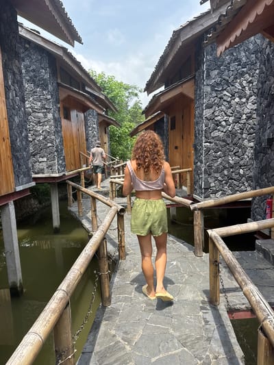 The image depicts an outdoor scene in Du Già Commune, Vietnam, on a bright, sunny day. The view is looking down a pathway or bridge that extends over a body of calm, murky green water.

In the foreground, a person with curly hair, wearing a light-colored tank top, green shorts, and yellow flat shoes, walks away from the camera, along the pathway. Further ahead, another person, wearing a cap, light t-shirt, and dark shorts, is also walking in the same direction.

The pathway itself is constructed from irregular dark grey flagstones. It is bordered on both sides by robust railings made of bamboo, with some sections connected by chains. Flanking the pathway are buildings characterized by their unique architecture. The lower parts of these structures feature facades of dark grey, rough-hewn stacked stones, while the upper sections are made of dark wood. The roofs exhibit intricate, scalloped wooden edges, adding a decorative element. Lush green trees are visible in the background and above the buildings, under a bright, clear sky. There is no visible text in the image.