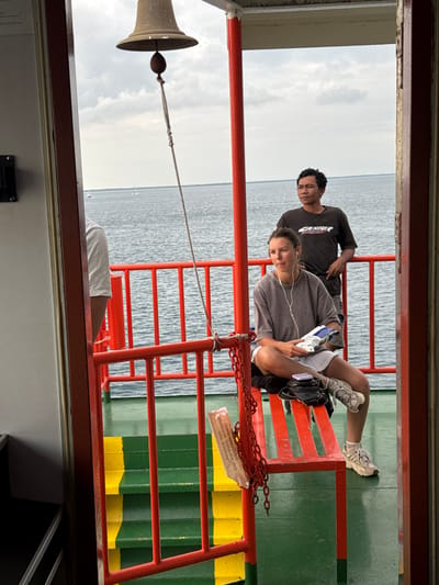 This image depicts an outdoor scene on a ferry at sea, possibly near Upt Tambak Sari, Indonesia. In the foreground, a woman with earbuds sits on a red bench, holding a package of snacks. She is wearing a grey t-shirt, white shorts, and athletic shoes. Behind her, a man in a dark t-shirt stands, looking towards the horizon. The ferry has green and yellow painted steps and red railings. A bell hangs from the ceiling in the upper left corner. The sea is calm, with a few boats visible in the distance under a cloudy sky.