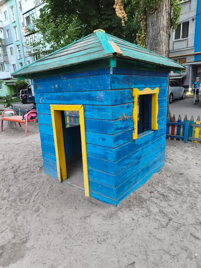 A bright blue wooden playhouse with a teal roof and yellow trim sits on a sandy playground. The playhouse has an open doorway and a square window. In the background, a colorful wooden fence, a silver car, and apartment buildings are visible. A person on a scooter rides past the fence. The scene appears to be a typical urban playground in Kropyvnytskyi, Ukraine.