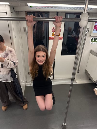 The image shows two people inside a subway car. In the foreground, a young woman with long, light brown hair is lying on her back, holding onto a metal pole with both hands. She is wearing a black dress and smiling at the camera. Her legs are bent, and she is positioned horizontally in the frame. Behind her, another person is visible, partially obscured, wearing a patterned sweater and dark pants. They are sitting down, with an umbrella resting beside them. The subway car interior is visible, including the floor, walls, and another metal pole. There are signs with Chinese text on the wall, indicating "当心夹手" (Beware of getting hands caught), "门灯闪烁" (Door lights flashing), and a route map. The overall impression is a playful and candid moment captured within a public transportation setting. The lighting suggests it is daytime. The visible text and the subway car design strongly suggest the location is a subway in China, specifically mentioned as Changning District.