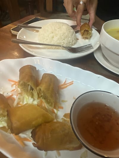 In an indoor restaurant setting in Tambon Ko Tao, Thailand, a close-up overhead shot displays a dining table laden with Thai cuisine. On a light brown wooden table, a white rectangular plate in the foreground holds approximately five fried spring rolls served on a bed of shredded cabbage and julienned carrots. Two of these spring rolls are cut in half, revealing a light green filling. Adjacent to this plate, on the lower right, is a small white bowl with a dark rim, containing a reddish-brown translucent dipping sauce with visible small red chili flakes.

In the midground, a white round plate features a dome-shaped mound of white rice, with a silver fork resting on its left side. Above this plate, a human hand, adorned with a silver ring on the ring finger, is seen holding a partially consumed fried spring roll, revealing its inner contents. To the right of the rice plate, a white bowl containing a yellow liquid, likely a soup or curry, is partially visible. A dark-colored smartphone lies on the table to the left of the rice plate. The background features blurred wooden surfaces, consistent with a restaurant environment.
