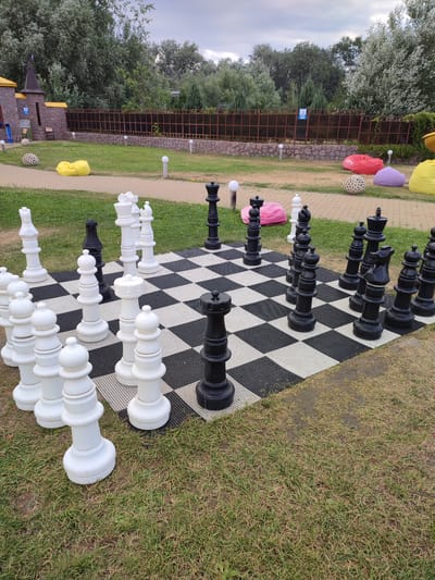The image shows a life-sized outdoor chess set on a checkered mat. The chess pieces are predominantly white and black, with several white pawns and a white queen in the foreground. Behind them, a black king and queen are visible, along with other black pieces. The chess set is placed on a grassy area, with a paved walkway visible in the background. Beyond the walkway, there is a low stone wall topped with a dark metal fence. In the distance, there are trees and a cloudy sky. To the left of the chess set, a small, castle-like structure with yellow turrets is partially visible. Scattered around the scene are several large, colorful beanbag chairs in yellow, pink, and purple, as well as decorative spherical objects. The overall setting appears to be a recreational area, possibly a park or a resort, in Shcherbani, Ukraine. The time of day seems to be daytime, with overcast weather indicated by the sky. No people are visible in the image.