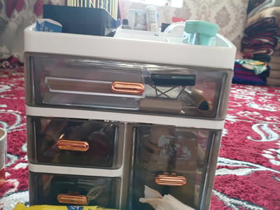 The image shows a white and clear plastic organizer with multiple drawers, filled with various cosmetic items. The organizer is placed on a red and white patterned carpet. In the background, there is a window with daylight filtering through and a stack of folded colorful fabrics. A small blue and white packet with text is visible in the top compartment of the organizer. The organizer has rose gold colored drawer pulls. The scene is indoors, likely in a residential setting, and appears to be in a room within the city of Uchkun, Kyrgyzstan. There are no people visible in the image.