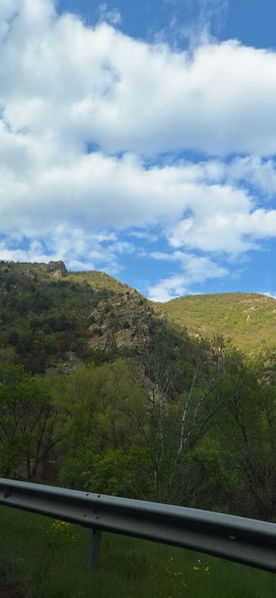 The image depicts a landscape with a sky filled with white cumulus clouds against a blue sky. A mountain or hillside with varying vegetation, including trees and some exposed rock faces, extends vertically through the frame. A metal guardrail is visible in the foreground on the right side. The location is identified as Mechkul, Bulgaria.