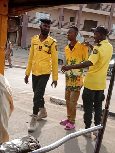 Three men stand on a street. One wears a yellow uniform with black accents, a black beret, and tan boots. Another wears a yellow palm tree shirt, tan pants, and pink sandals. The third wears a yellow shirt, black cap, and black pants. A building is in the background. A person walks by. A car is in the background.