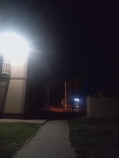 An outdoor nighttime scene in Znamensk, Russia, features a building with a brick facade illuminated by a bright light source in the upper left corner. A metal railing is visible on the side of the building. A paved pathway leads away from the building into the darkness. To the right of the pathway is a grassy area. Further in the background, another building with a dark roof and a light-colored wall is visible, along with a street light. The sky is dark and clear.