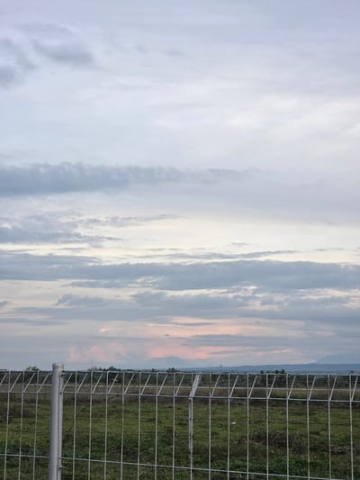 The image shows a metal fence running vertically along the right side of the frame, in what seems to be a rural area near Pintomakmur, Indonesia. Beyond the fence is a field of green grass and low vegetation. The background features a horizon line with a faint outline of hills or distant mountains. The sky occupies most of the frame, displaying streaks of light clouds with hues of grey, blue, and hints of orange possibly indicating sunrise or sunset. The image is taken from ground level and is slightly angled.