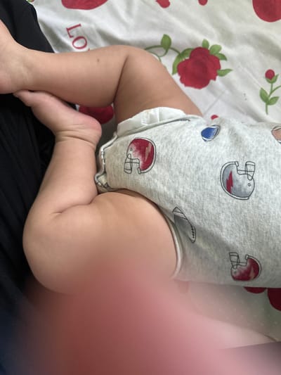 The image shows a close-up of a baby's legs. The baby is lying on a bed with a patterned sheet. The sheet has images of strawberries and roses. The baby is wearing a grey onesie with a pattern of football helmets. The baby's left foot is resting on its right leg. The image is taken from a high angle, looking down at the baby's legs. There is a blurry pink area in the foreground which is likely a finger obstructing the camera lens. The image does not provide any information about the location in the city of Тайгараев, Kyrgyzstan.