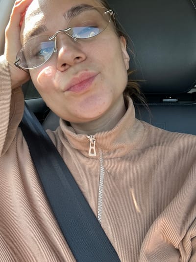 The image shows a close-up, selfie-style shot of a person wearing rimless glasses and a tan corduroy jacket with a zipper. The person's left hand is resting on their forehead. A black seatbelt is visible across the person's chest and shoulder. The background appears to be the interior of a vehicle, with a dark headrest visible. The lighting suggests it is daytime. There are no discernible landmarks or indicators to place the location in Uva, Russia.